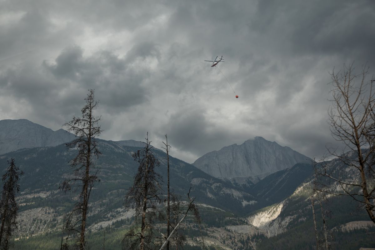 A helicopter buckets water onto smouldering fires outside of Jasper, Alta., on Friday July 26, 2024. Wildfires encroaching into the townsite of Jasper forced an evacuation of the national park and have destroyed over 300 of the town's approximately 1100 structures, mainly impacting residential areas.