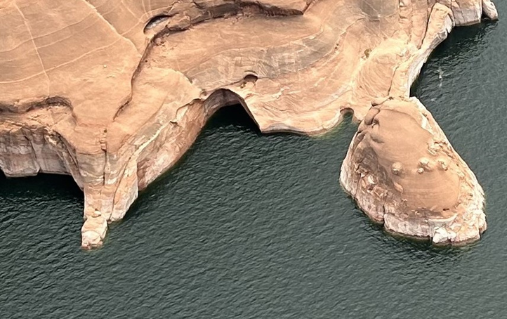 Orange, sandstone rock formation juts out of dark blue water, showing the aftermath of the 'Double Arch' collapse.