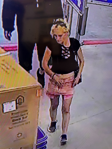 Caucasian female walking into a store. The female is wearing a pink shirt, black top, black and white runners, with sunglasses on her head.