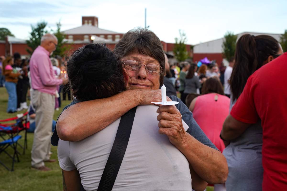 Two women embrace. One woman is holding a candle. There is a crowd in the back of the photo.