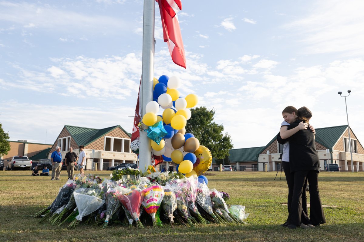 Two students hug beside a flagpole surrounded by flowers in memorial.