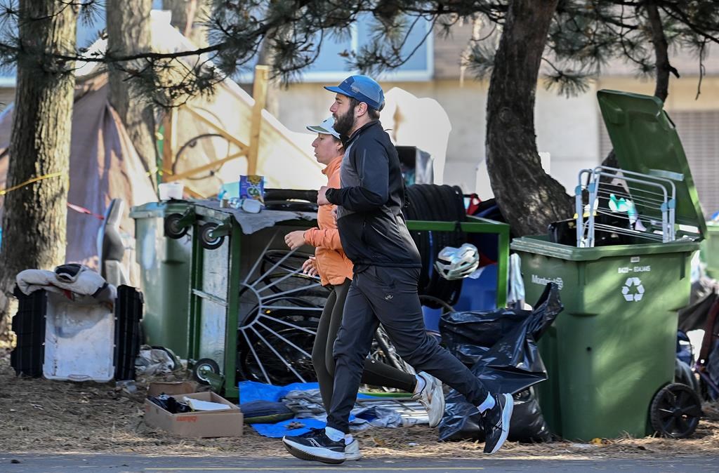 People jog by a homeless encampment on Notre-Dame Street in the east end of Montreal, Friday, Oct. 25, 2024. 