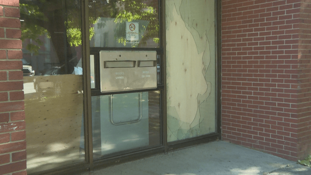 The side of the library. The picture shows the metal book chute, glass panes, and the broken glass on the right pane. It has a wooded board behind it, and there's a brick wall to the right of the glass.