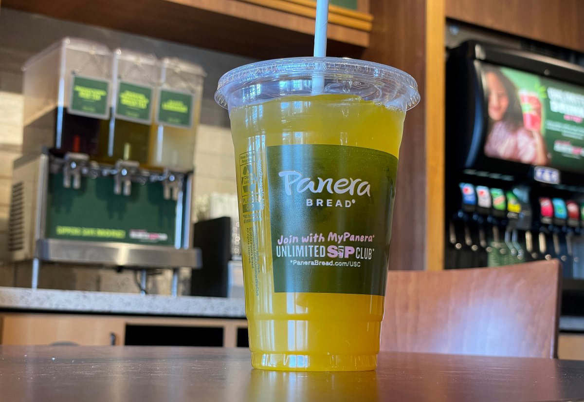 Yellow liquid in a Panera Bread to-go plastic cup. Drink dispensers are seen in the background.