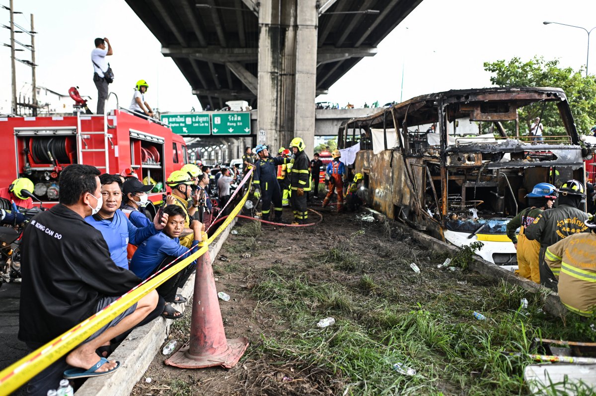 Rescuers and firefighters stand behind caution tape, beside a burned shell of a double-decker bus.