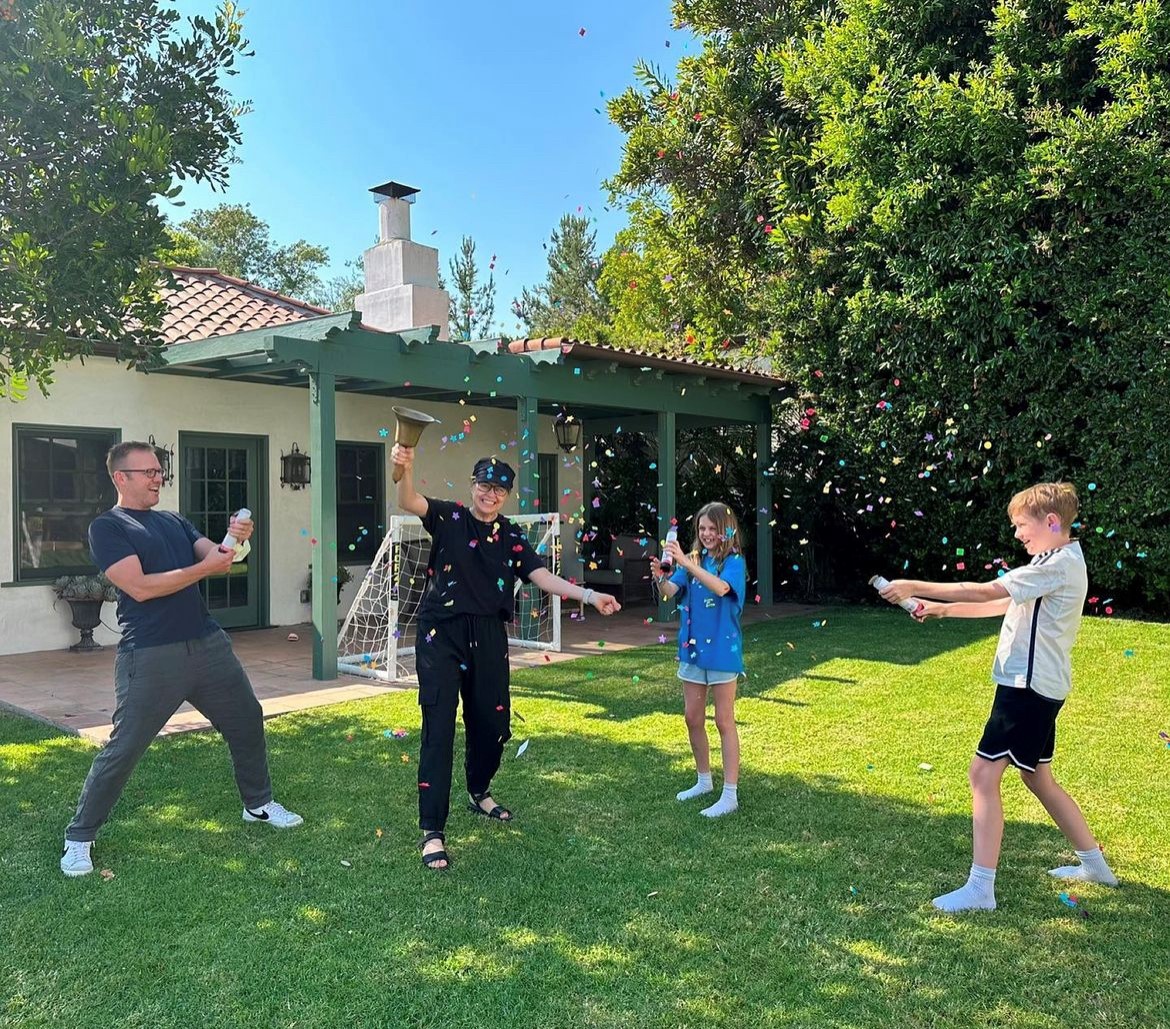 Jenna Fischer rings a bell in a backyard beside her husband, Lee, and their two children, who are throwing confetti.