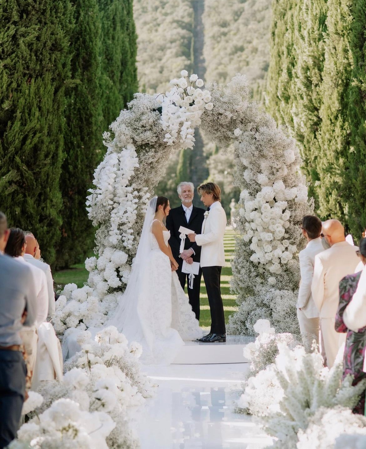 Millie Bobby Brown and Jake Bongiovi exchange vows in front of Matthew Modine under a floral arch.