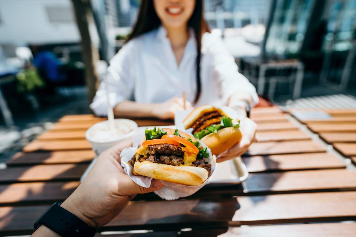 A happy young couple eating burgers and fries outside.