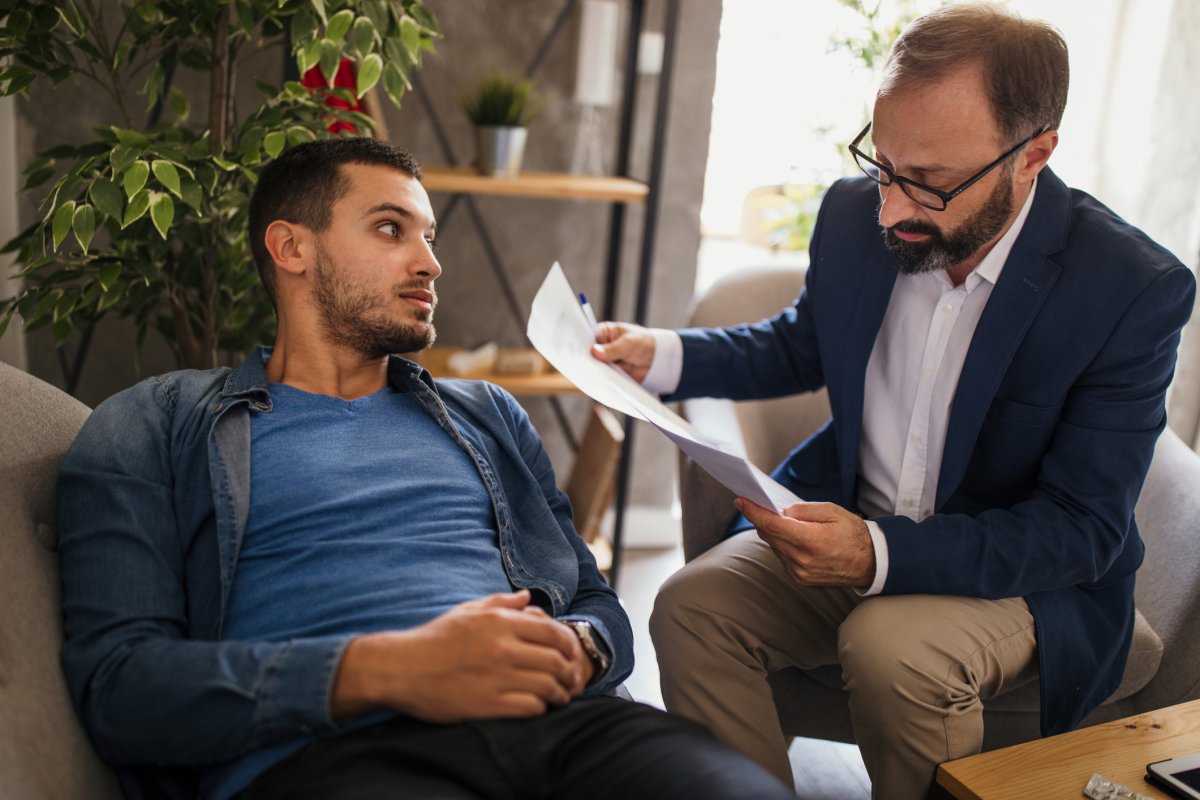 A man examines documents while another man lays on a couch.