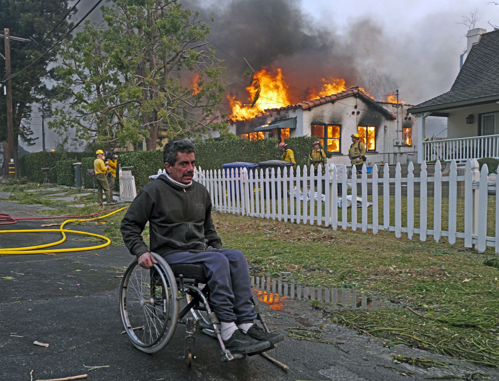 A man in a wheelchair goes past a house on fire from the Eaton Fire in the Altadena neighborhood on January 08, 2025 in Pasadena, Calif.