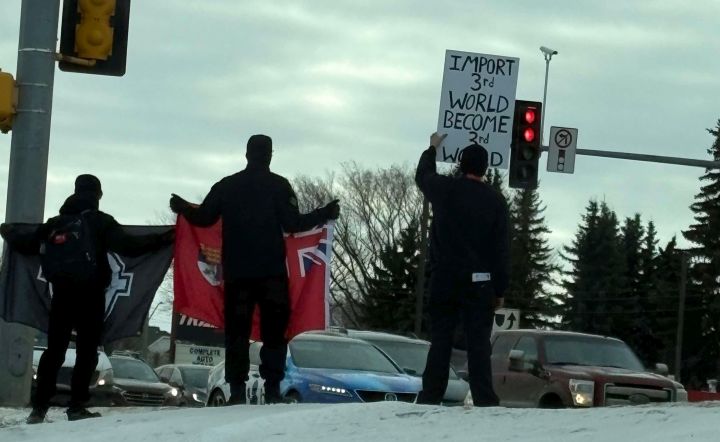 Three people are seen standing by a busy road in St. Albert, Alta., on Jan. 25, 2025 holding signs and flags.