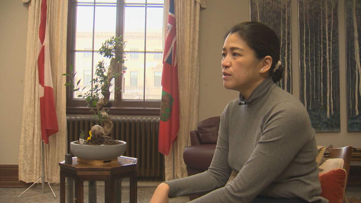 A woman sits in a windowed office. She is wearing a grey sweater. there are flags flanking the office's window and a plant on a stand sits in front of the window.