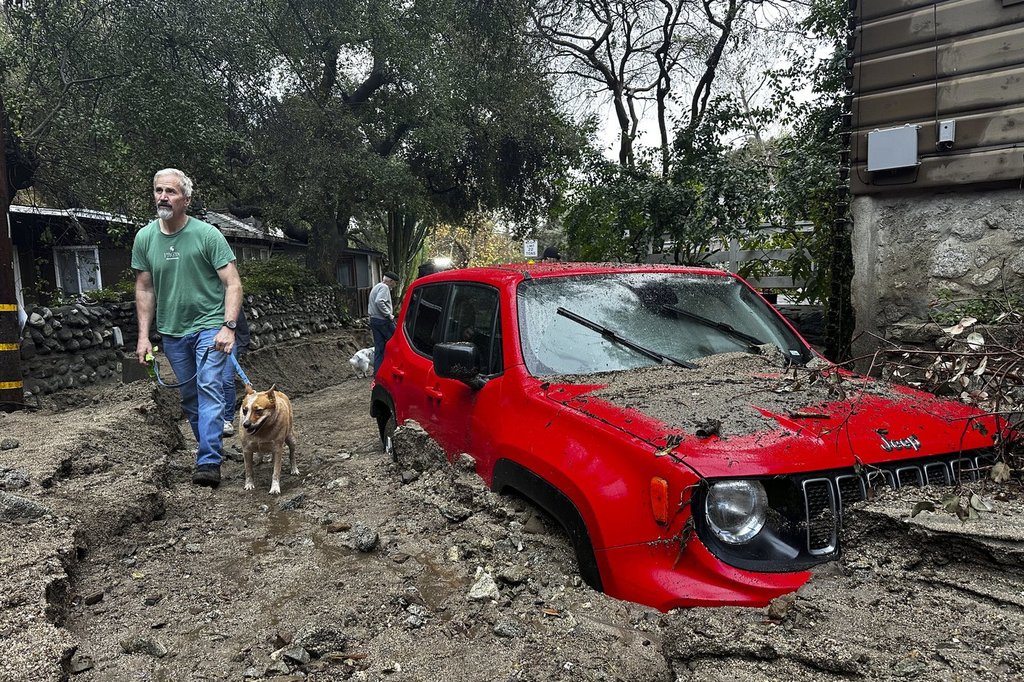 A resident and their dog walk past a vehicle partially submerged in mud after a storm Friday, Feb. 14, 2025, in Sierra Madre, Calif.