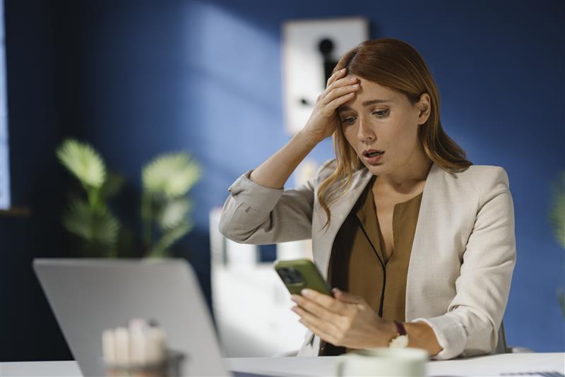 A businesswoman reads a shocking text message on her phone while sitting at her desk.