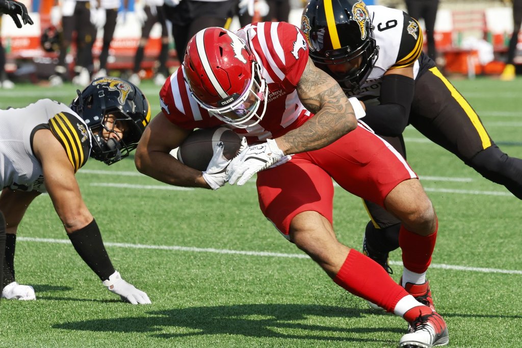 Calgary Stampeders' Reggie Begelton, centre, is tackled by Hamilton Tiger-Cats' Jonathan Moxey, right, during first half CFL football action in Calgary, on Saturday June 7, 2025.