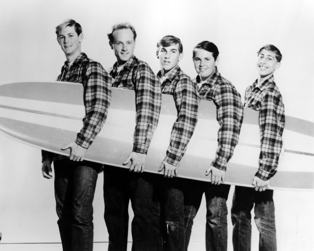 L-R: The Beach Boys’ Brian Wilson, Mike Love, Dennis Wilson, Carl Wilson, David Marks pose with surfboard early in their careers.