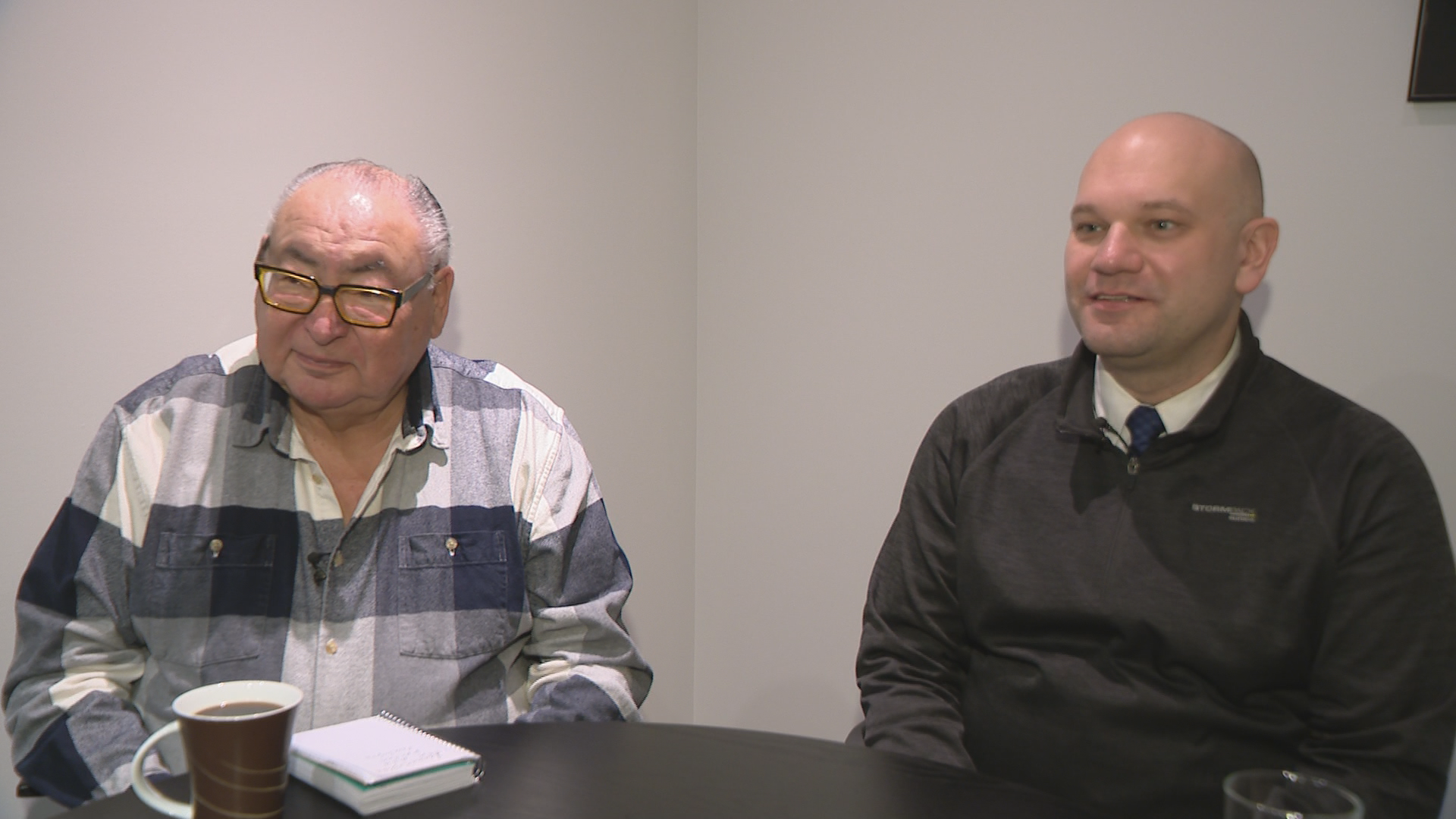 Two men sit at a table. The one on the left is wearing a white and black plaid shirt while the one on the right is wearing black.