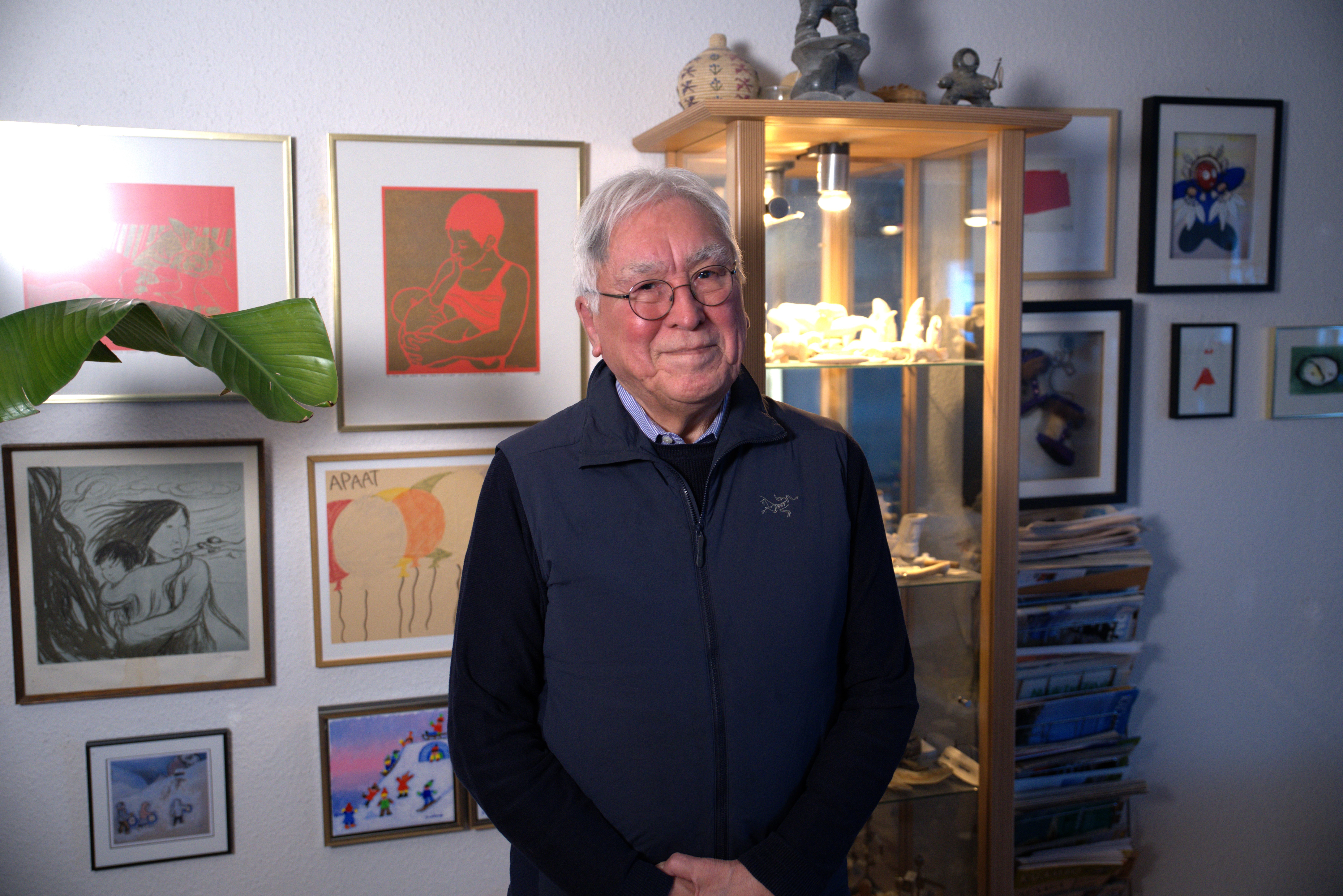 A white-haired man in a blue vest and black sweater stands in front of a wall of filled with artworks and a shelf of Inuit carvings