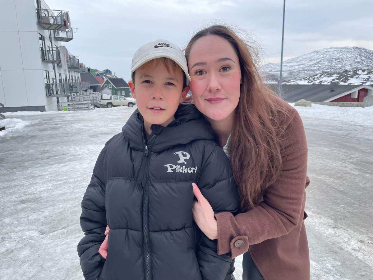A woman with long brown hair wearing a brown coats stands behind a boy in a black puffer vest and white cap. They are outside on a snowy road.