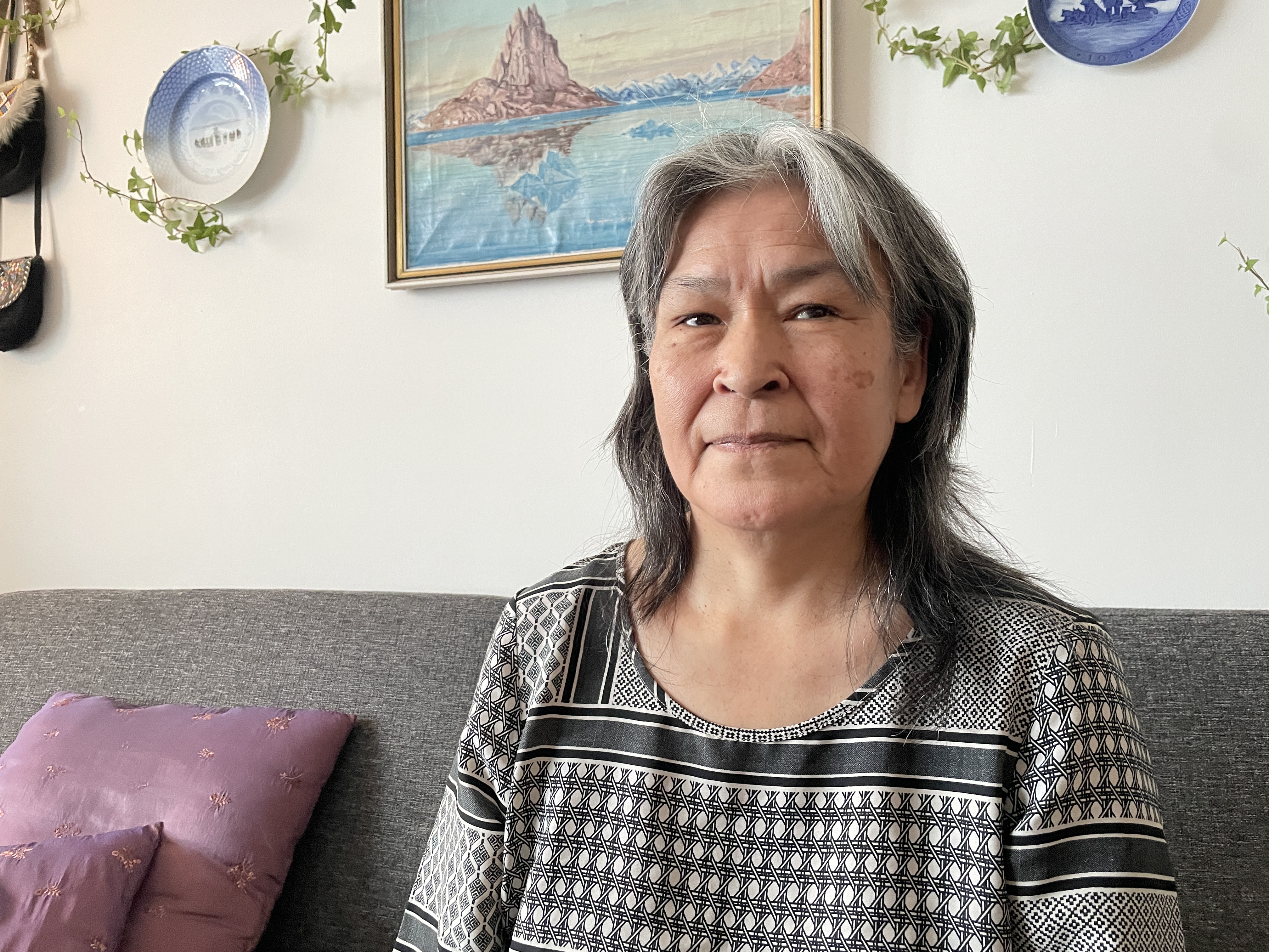 An Inuk woman with long greying in a grey and white striped T-shirt sits on a couch and looks worried into the camera.