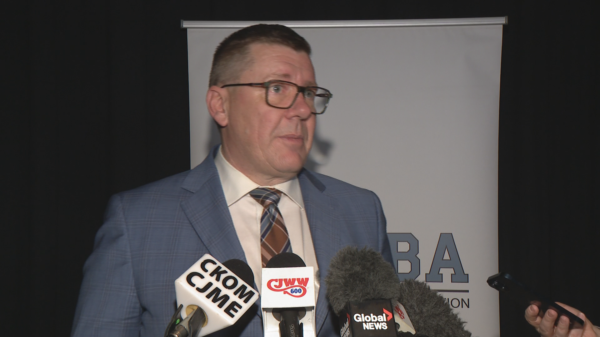Premier Scott Moe speaks to reporters in front of a white banner and black backdrop.