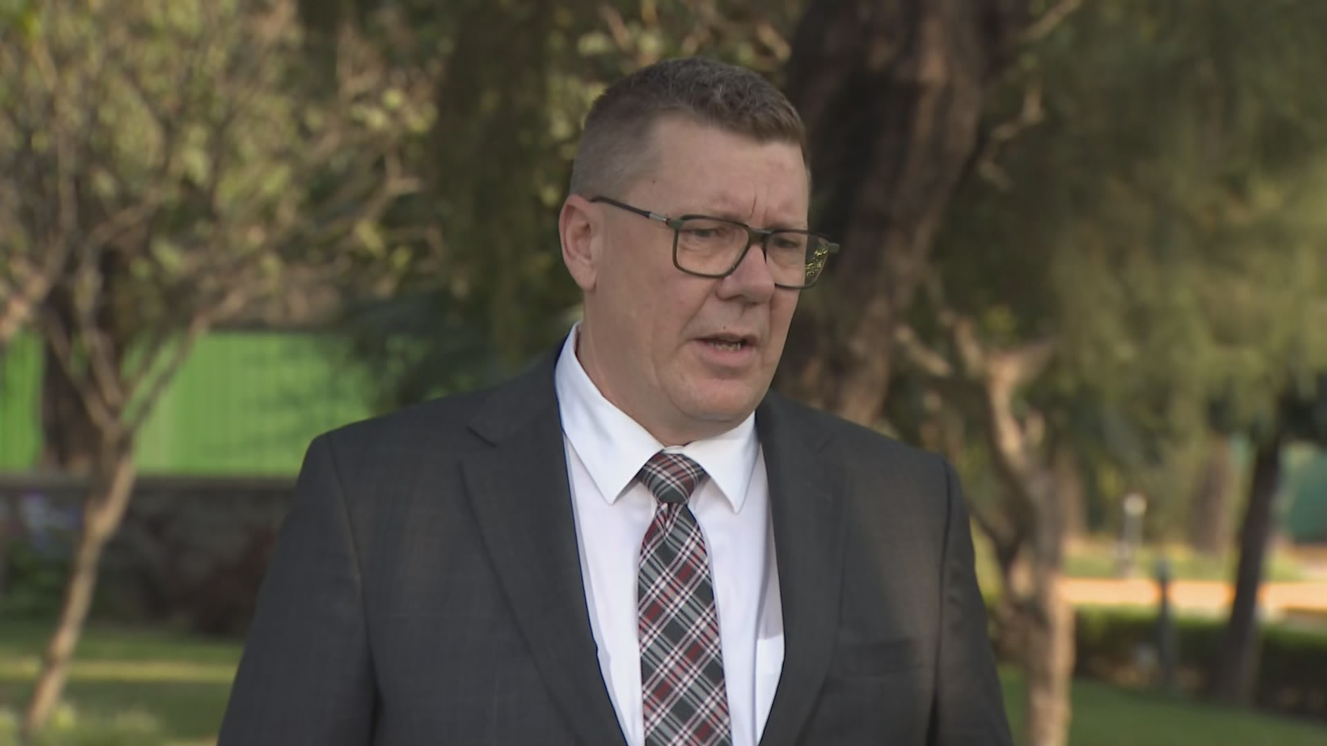 Premier Scott Moe stands in a suit and tie in a park in New Delhi with trees behind him and other greenery.