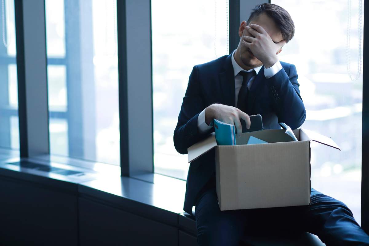 A fired businessman hides his face and sits near a window with his office supplies.
