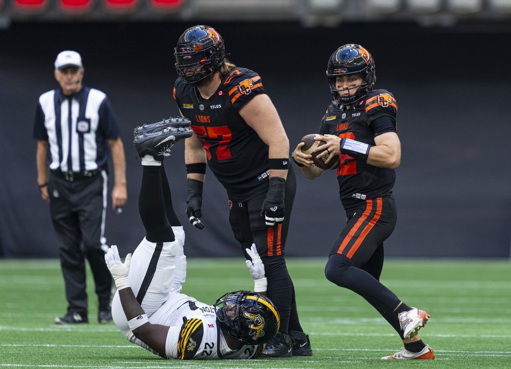 Four people, three in sports uniforms and one in a referee uniform, are on a green field.