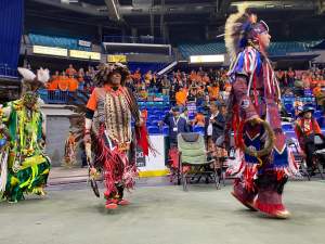 Men wearing regalia of feathers and colours walk inside an arena.