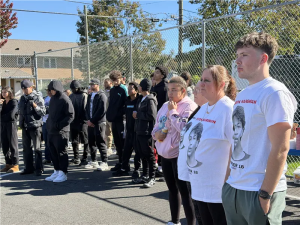 Theresa Gray stands with one of Devon's friends. They are both wearing white T-shirts with his face on them that read "Forever 16." in the background a crow of people wearing dark clothing stand along the basketball court's fence.